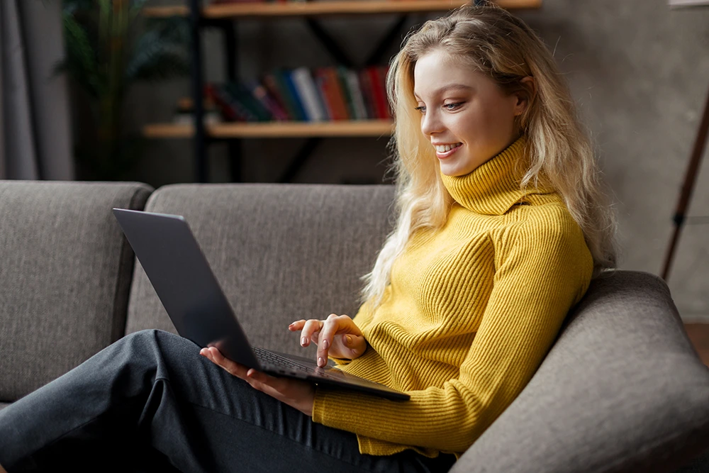 Woman wearing yellow turtle neck sitting on couch looking at her laptop during online therapy session with Zoom Therapy